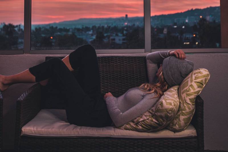 Woman wearing beanie hat lounges on wicker bench overlooking island at dusk